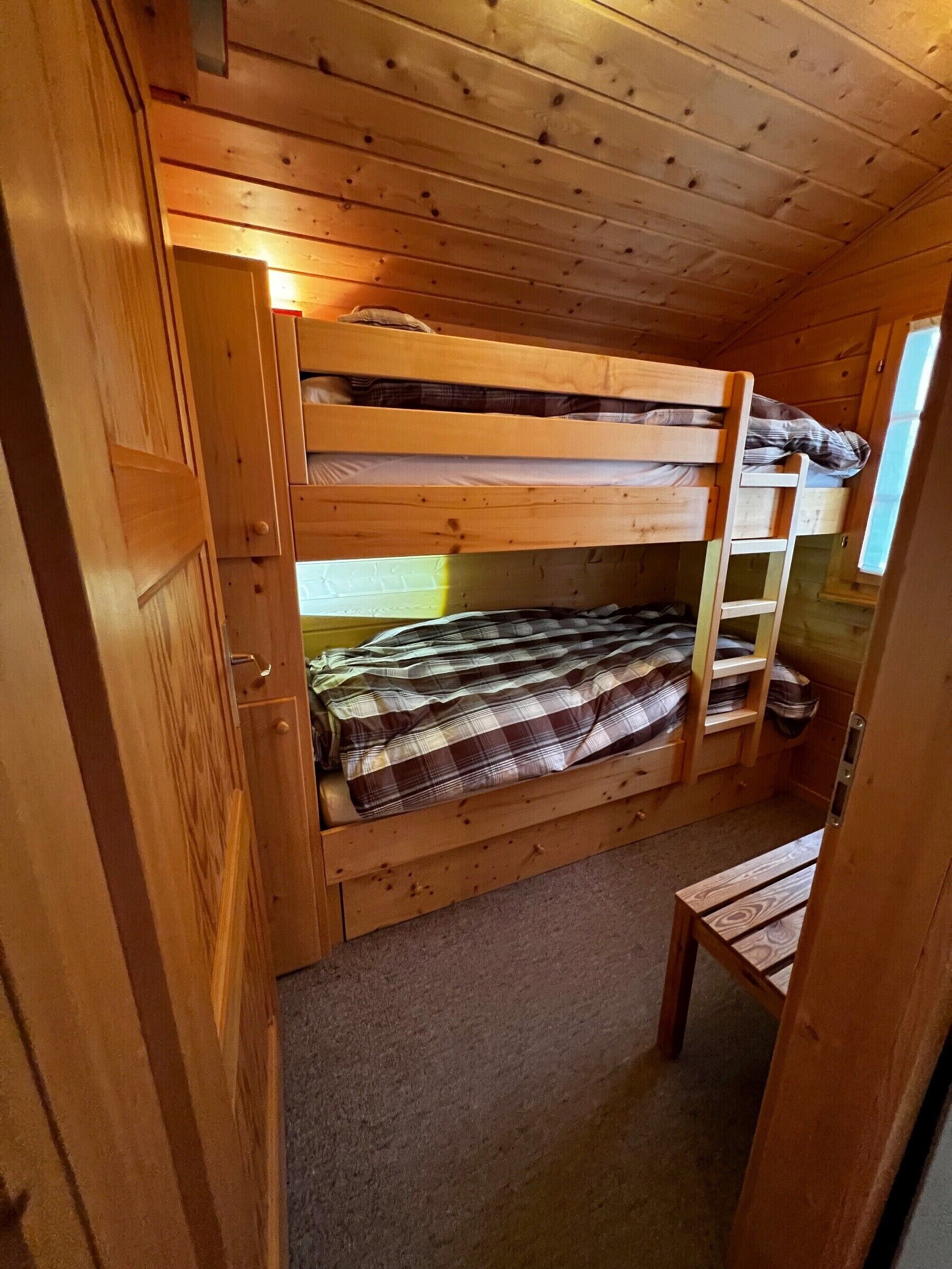 Habitación con literas, techo de madera, una ventana pequeña y un banco de madera.
