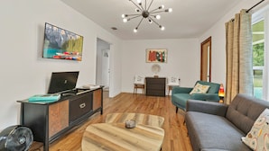Una sala de estar moderna con piso de madera, un sofá gris, un sillón verde azulado, una mesa de centro de madera, un mueble de entretenimiento negro, un televisor de pantalla plana y una lámpara de techo original.