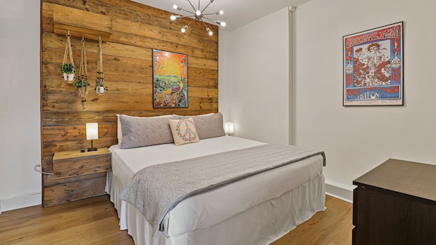 Un dormitorio con una pared de acento de madera, una cama con cubrecama blanco, una mesita de noche con lámpara y una obra de arte en la pared.