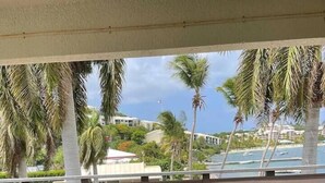 Ein überdachter Außenbereich mit Korbmöbeln und einem kleinen Tisch, mit Blick auf einen Strand mit Palmen und Gebäuden im Hintergrund.