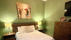Un dormitorio con una cama, dos mesitas de noche con lámparas, un aparador, un cuadro en la pared y un ventilador de techo.