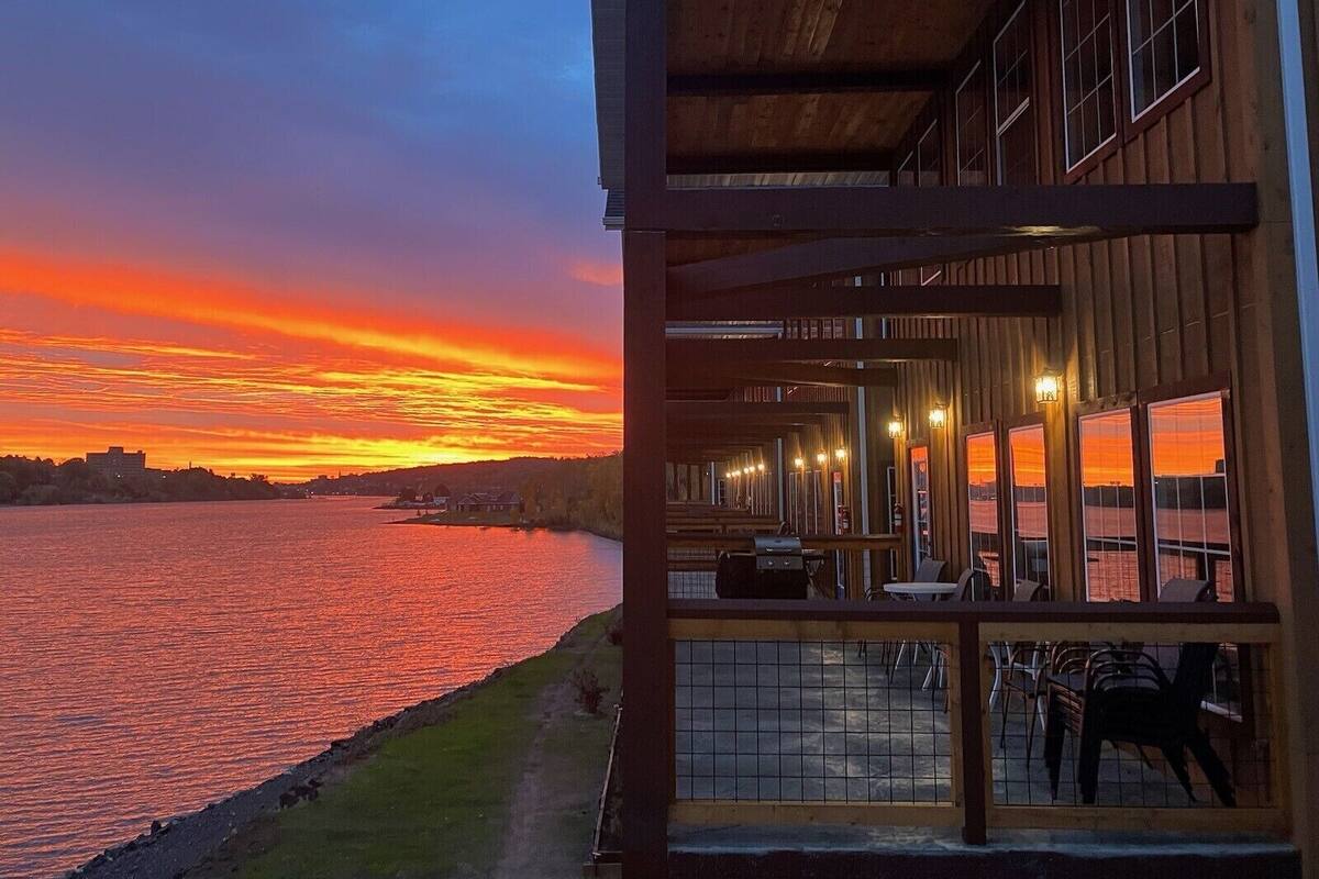 Een gebouw aan het water met een overdekt terras, grote ramen en een uitzicht op de ondergaande zon.