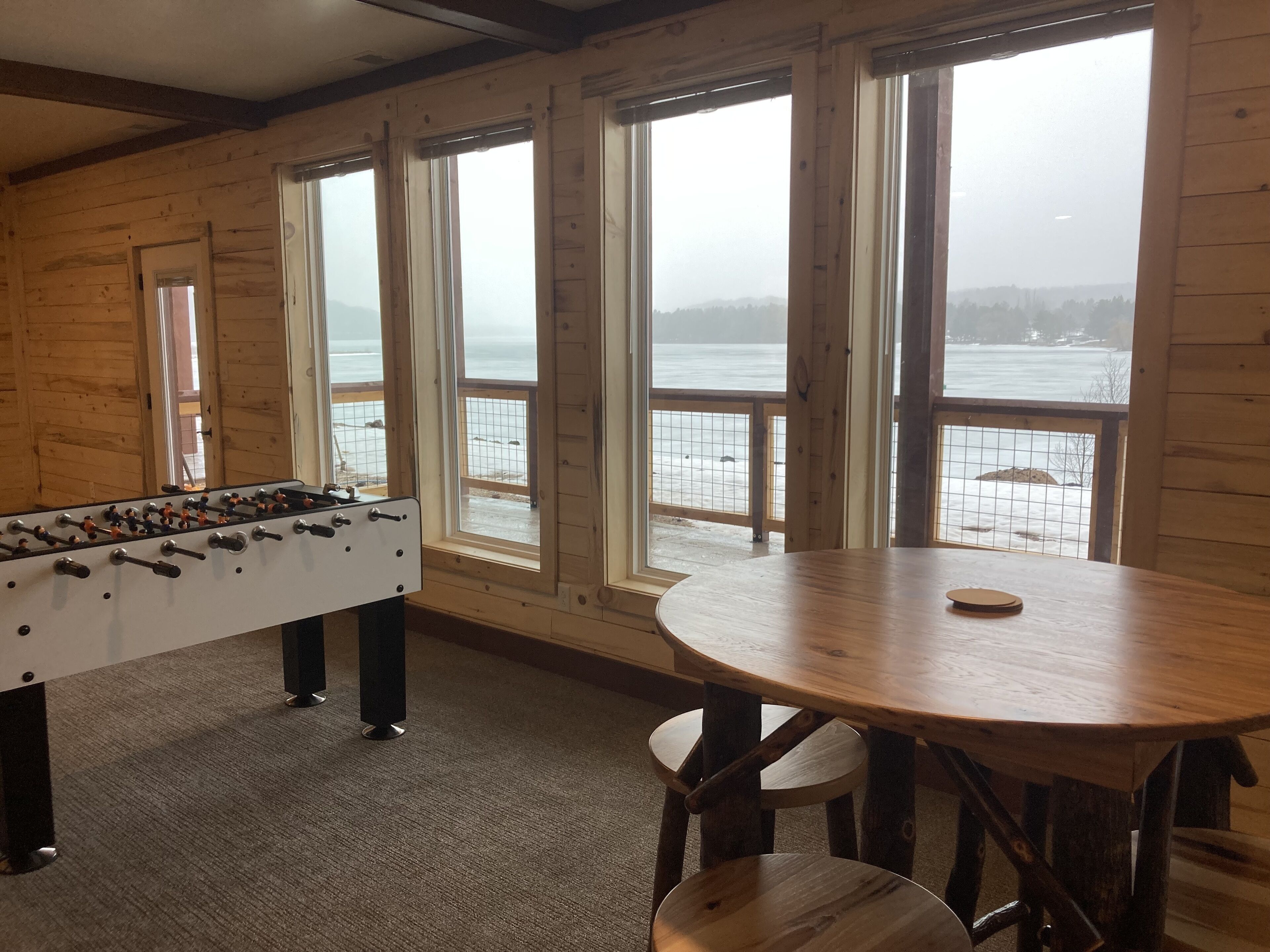 Ein Zimmer mit Holzboden, einer Tischfußballanlage und einem runden Tisch mit Stühlen. Große Fenster bieten Blick auf eine verschneite Landschaft und ein Gewässer.