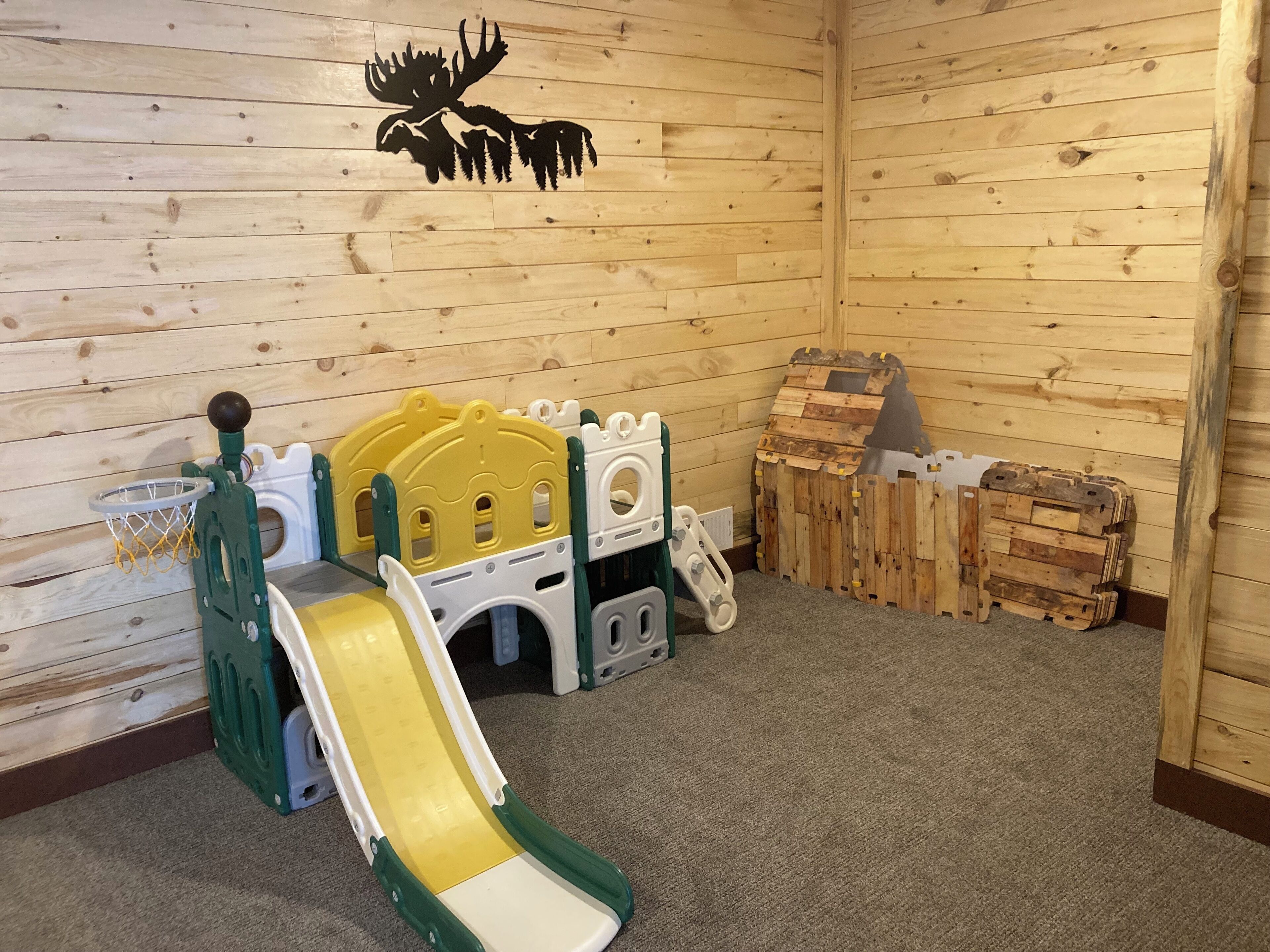 Een houten speelruimte met een glijbaan, een klein basketbalnet en een muursticker van een eland.
