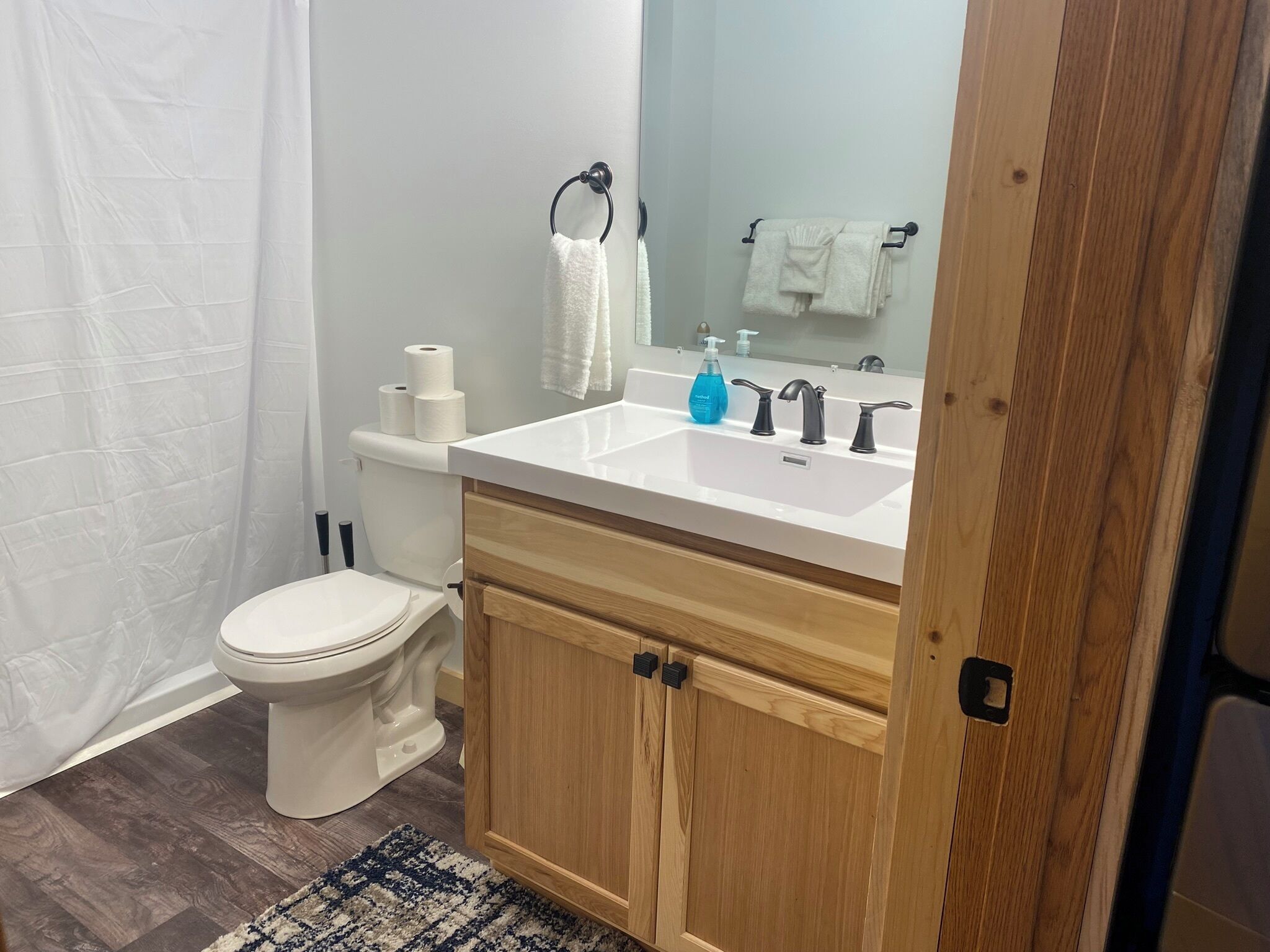 A bathroom with a white toilet, a white sink with a mirror above it, and a wooden cabinet below. There are two rolls of toilet paper on the toilet tank, a blue bottle of hand soap on the sink, and a white towel hanging on a towel rack.