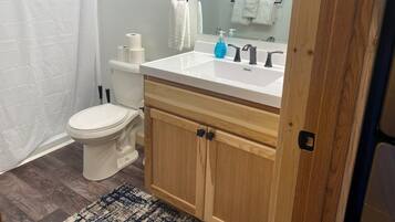 A bathroom with a white toilet, a white sink with a mirror above it, and a wooden cabinet below. There are two rolls of toilet paper on the toilet tank, a blue bottle of hand soap on the sink, and a white towel hanging on a towel rack.