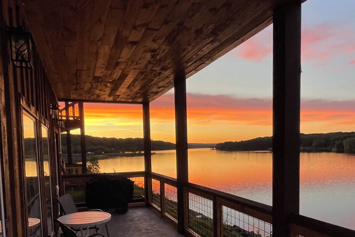 Een overdekt terras met uitzicht op een zonsondergang boven een meer, met buitenmeubels en een balustrade.