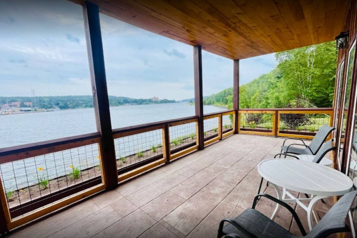Een overdekt buitengebied met uitzicht op een rivier en heuvels. Er staan stoelen en een tafel.