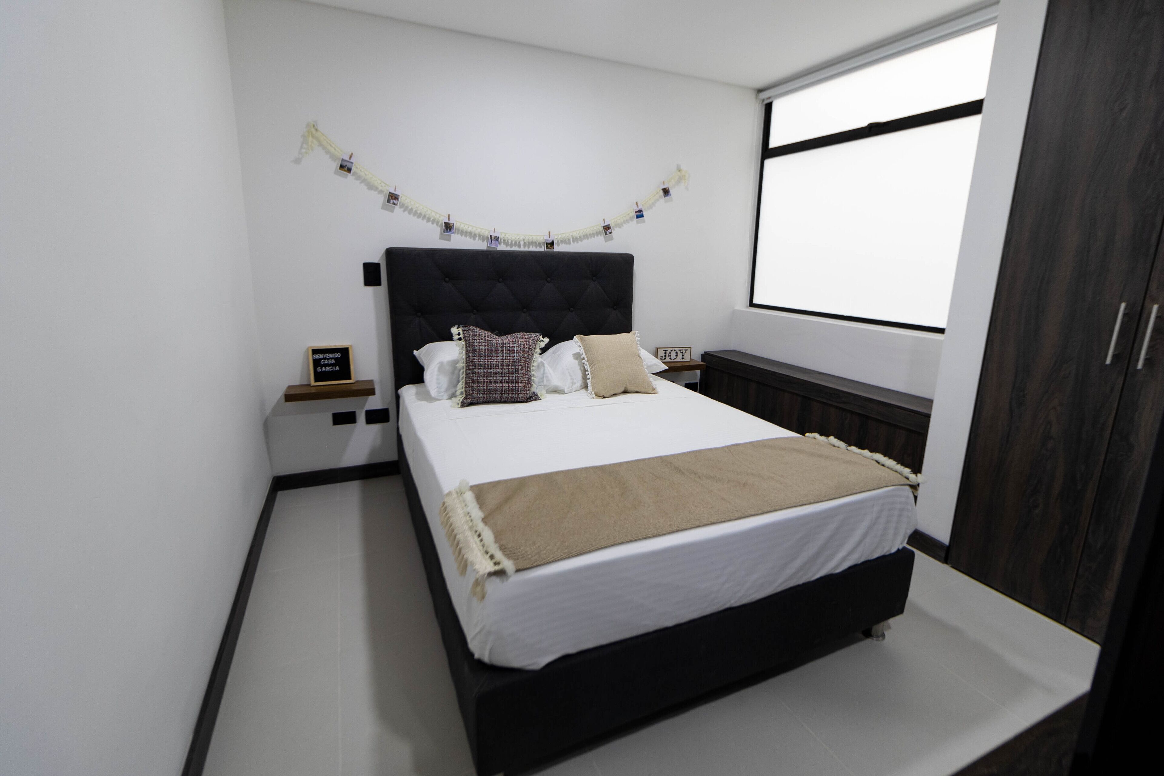 Un dormitorio ordenado con cama, mesitas de noche, un armario de madera oscura y una ventana con vistas.