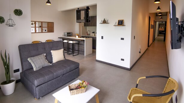 A modern living room with a grey sofa, a white coffee table, and a wicker chair. A kitchen area is visible in the background with a dining table and bar stools. - Casa García Rent Home (Guatapé)