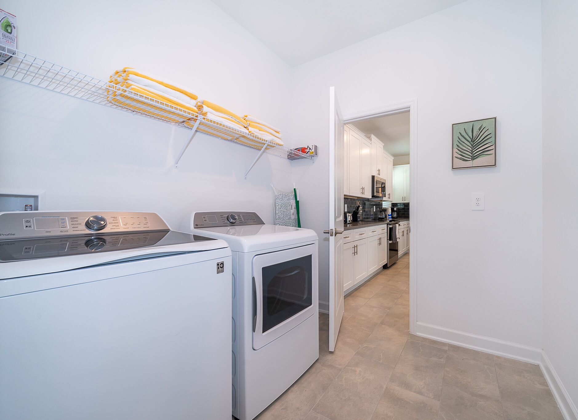 A laundry area with stacked laundry, a washing machine, and a dryer. There is a kitchen visible in the background with white cabinets and a microwave.