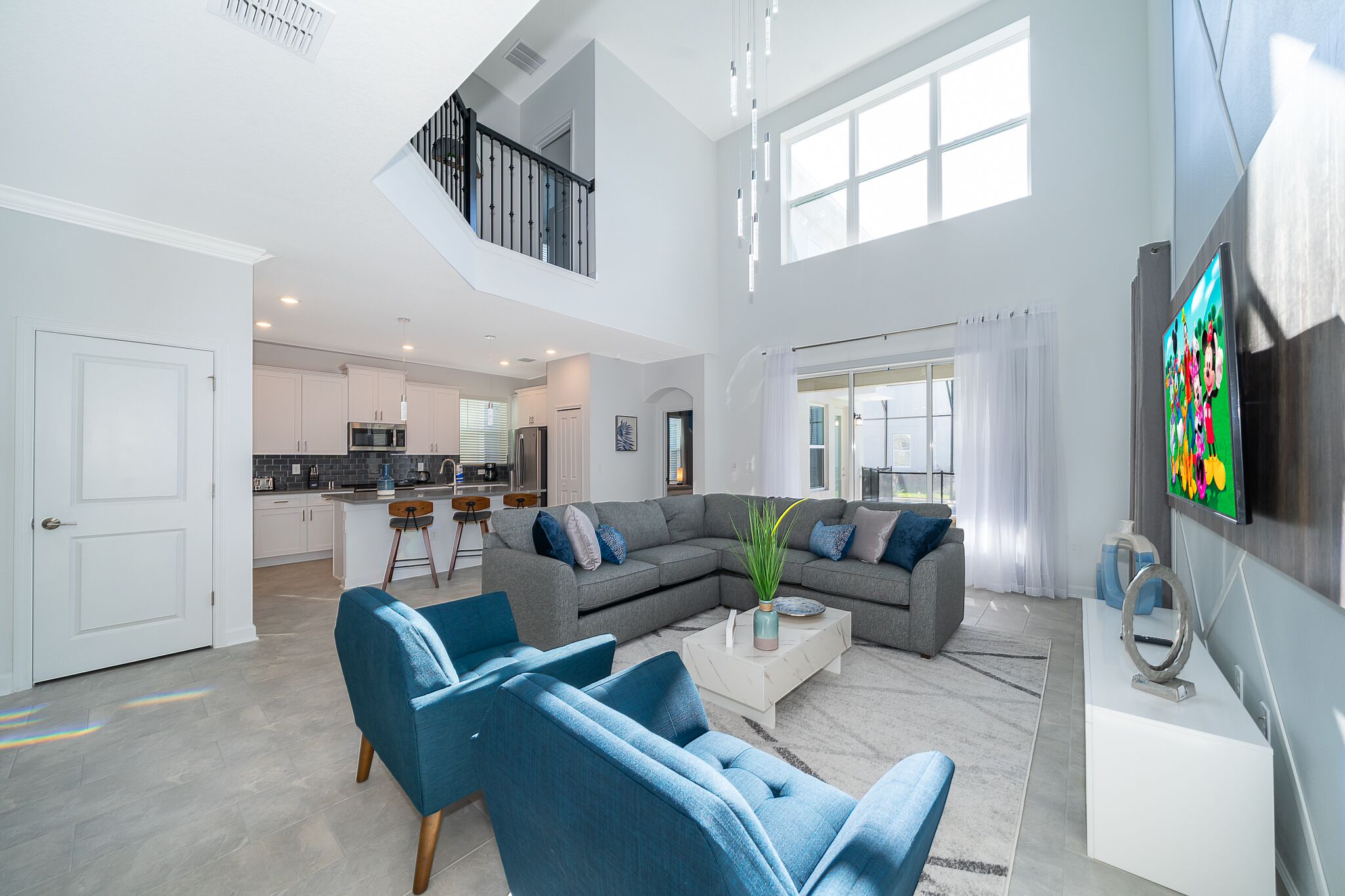 A spacious living area with a large gray sectional sofa, blue armchairs, and a coffee table. A kitchen is visible in the background, and a staircase leads to an upper level.