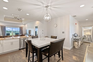 A spacious kitchen with a central island, a dining area with a white table and dark chairs, and a view of the outdoors through large windows. - Seven Eagles Lavish: Water Park, Golf, Near Disney (Kissimmee)