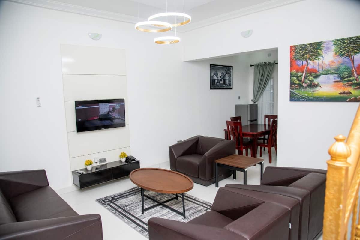 A modern living room with a flat-screen TV, brown leather sofas, a wooden coffee table, and a dining area with a wooden table and chairs.