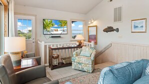 Ein Wohnzimmer mit Fernseher, einer Couch, einem Sessel, einem kleinen Tisch und einem Fenster mit Blick auf den Strand.