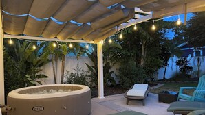A covered outdoor area with a hot tub, lounge chairs, and string lights.