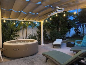 A covered outdoor area with a hot tub, lounge chairs, and string lights.
