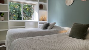 A bedroom with two beds, a window with a view of greenery, and a wall with woven decor.