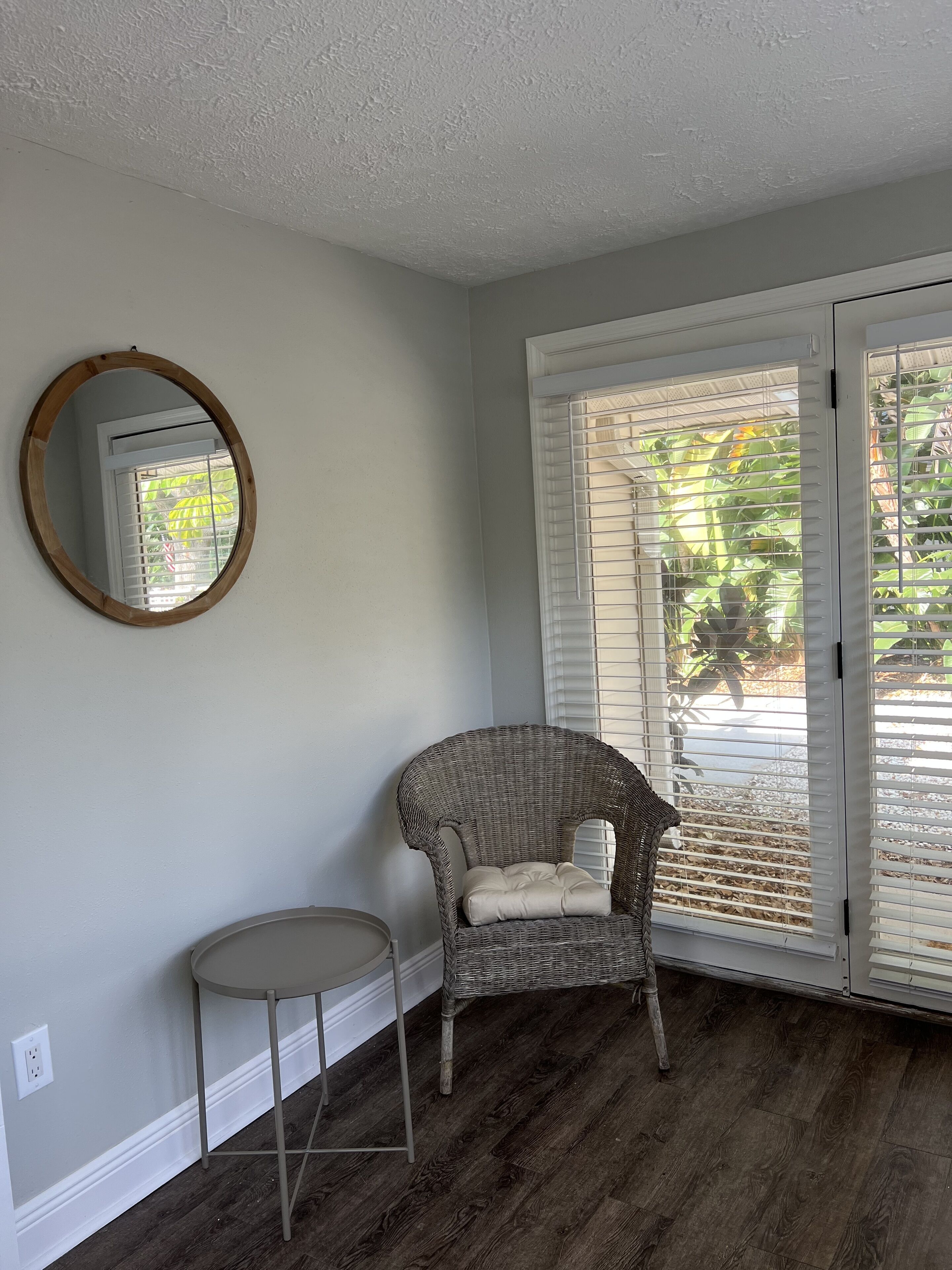 Una habitación con una silla de mimbre, una mesita redonda, un espejo circular y una puerta corredera de vidrio con persianas.