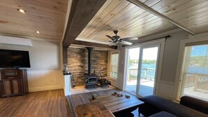 A cozy living room with a stone fireplace, a ceiling fan, and a large window overlooking a deck. - Bespoke Lake Cottage (Kings County)