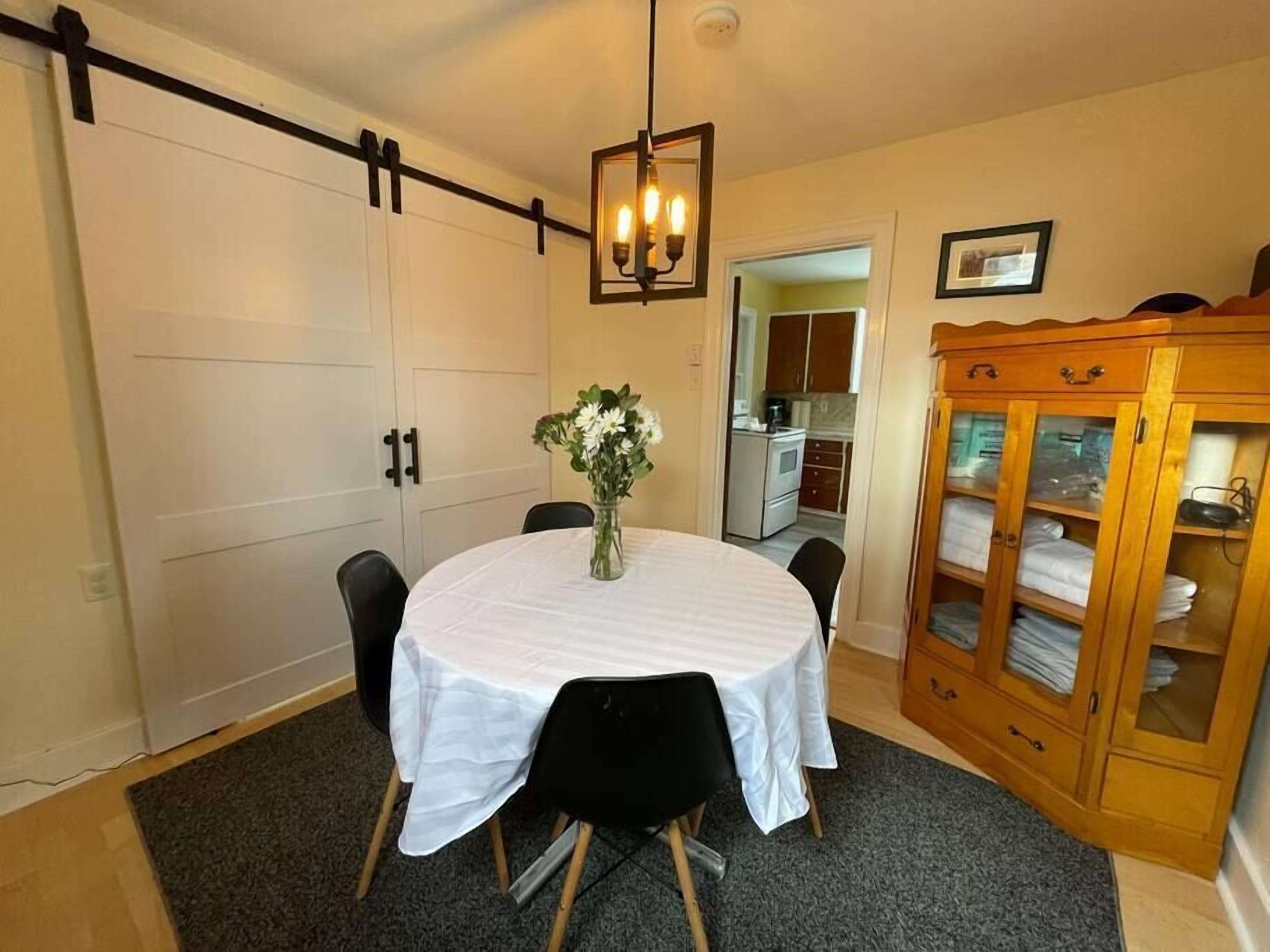 A dining area with a round table covered in a white tablecloth, surrounded by black chairs. There is a vase with flowers on the table. A wooden cabinet with glass doors is to the right.