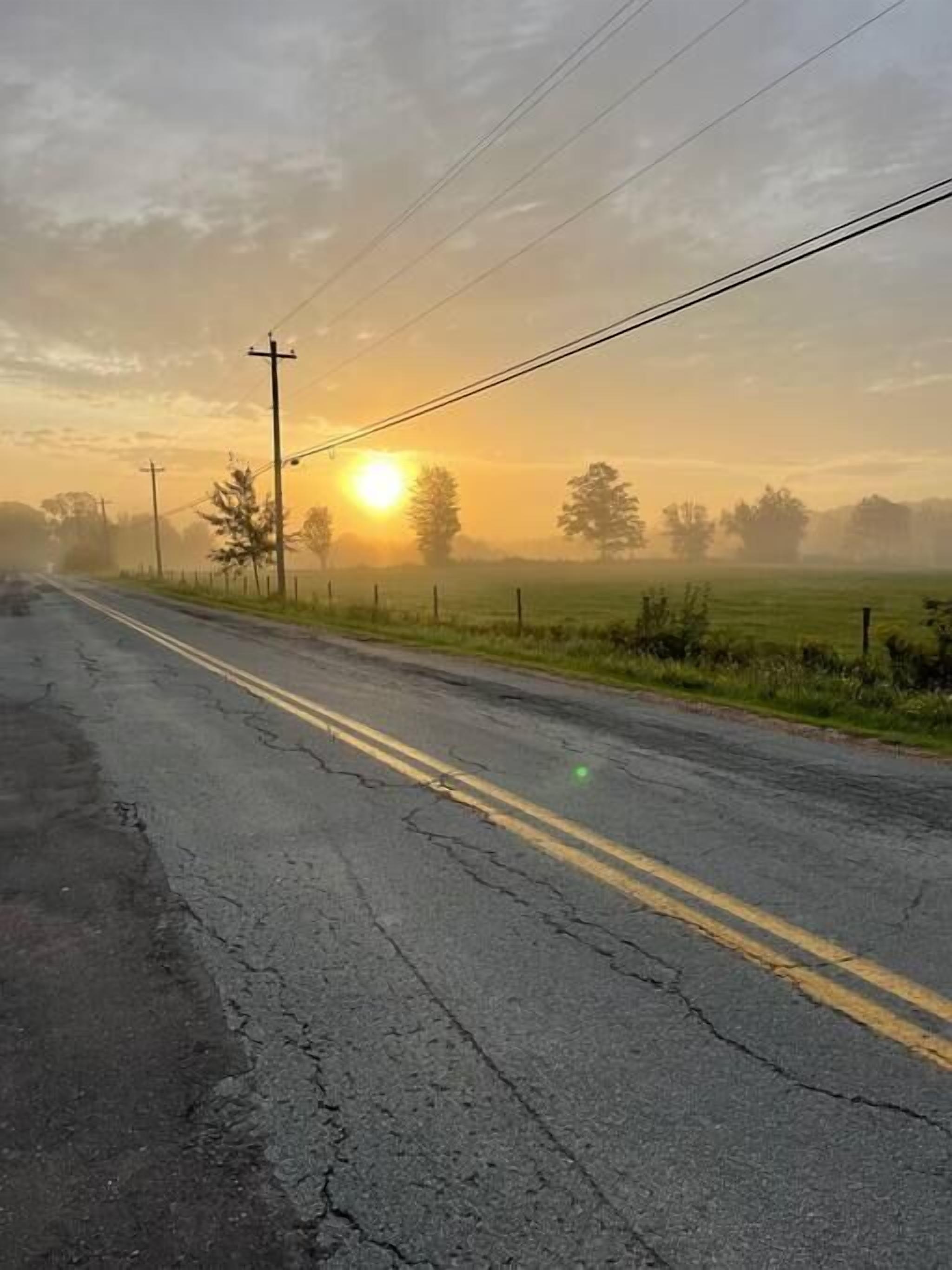 A rural road with a sunrise in the background, power lines, and a fence.
