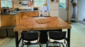 Une cuisine avec des meubles en bois, un îlot central doté de chaises, des ustensiles de cuisine suspendus et une fenêtre donnant sur un paysage verdoyant.