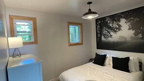 Une chambre à coucher avec un lit, une table de chevet, deux fenêtres et un tableau au mur.