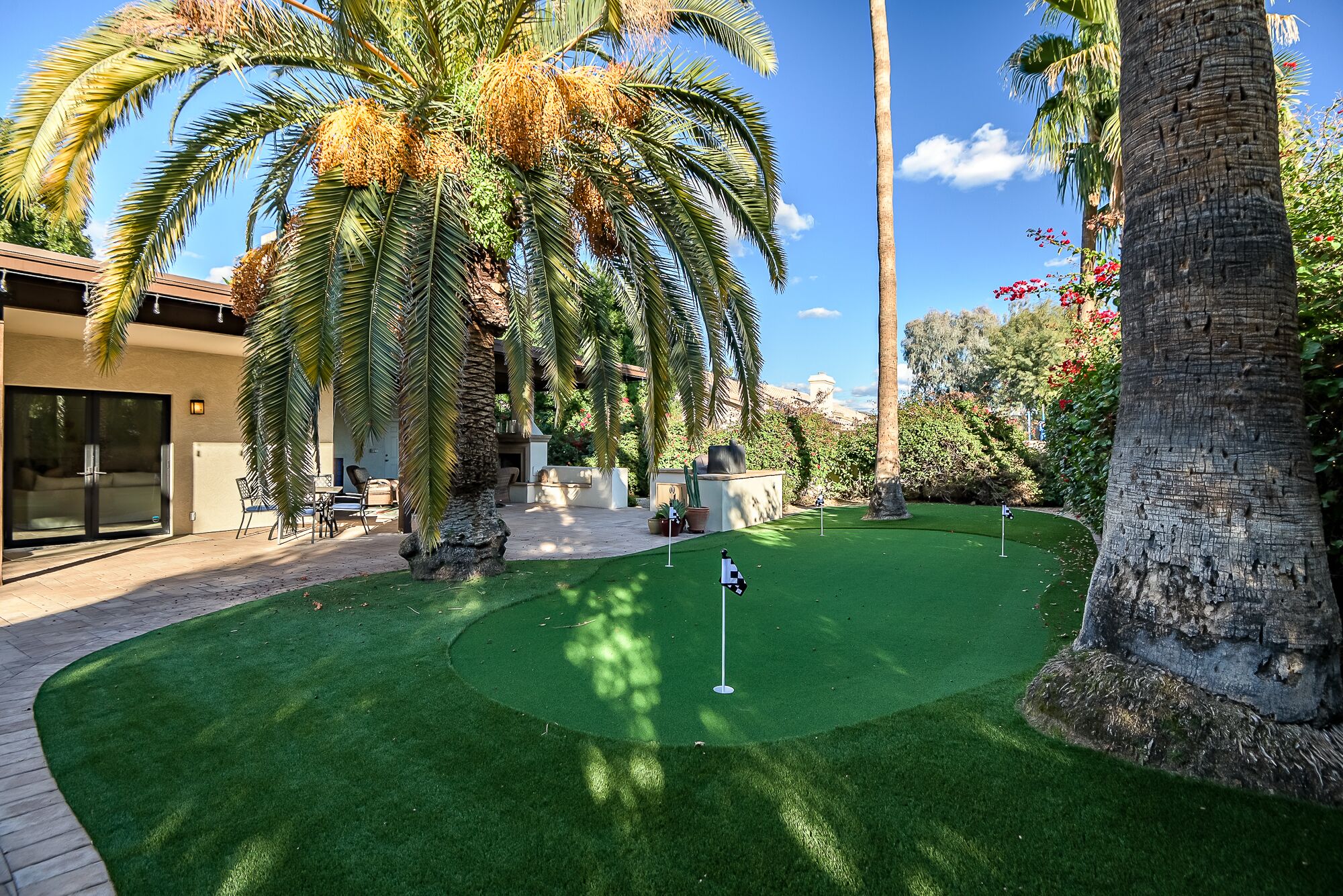 A well-maintained artificial golf course with a putting green, surrounded by palm trees and a building with a sliding glass door.