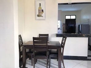 Un comedor con una mesa y cuatro sillas, un cuadro enmarcado en la pared, y una cocina visible a través de un vano abierto.