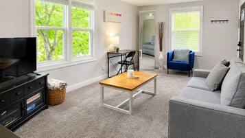 Un salon comprenant un téléviseur, une table basse, un canapé et un fauteuil. On y trouve une fenêtre avec des stores, une lampe et un vase avec des plantes.