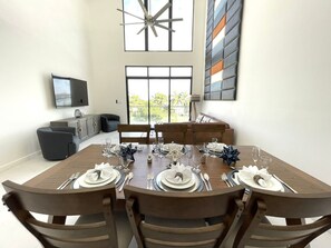 Uma área de refeições com uma mesa grande posta para quatro pessoas, cercada por cadeiras de madeira. Um ventilador de teto moderno e uma janela grande com vista para o verde são visíveis.