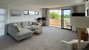 A modern living room with a grey sectional sofa, a coffee table, and a TV stand. There is a large window with curtains, a balcony, and an air conditioning unit on the wall. - Little Break Tahunanui - 5 minutes from beach (Nelson)