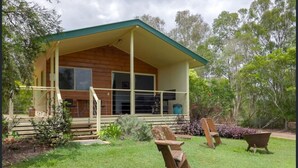 Une maison en bois avec un porche couvert, une porte d’entrée et un petit jardin comprenant une pelouse, un banc et une brouette.