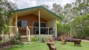 Ein Holzhaus mit überdachter Veranda, einer Haustür und einem kleinen Garten mit Rasen, einer Bank und einem Schubkarren.