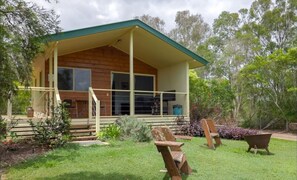 Une maison en bois avec un porche couvert, une porte d’entrée et un petit jardin comprenant une pelouse, un banc et une brouette.