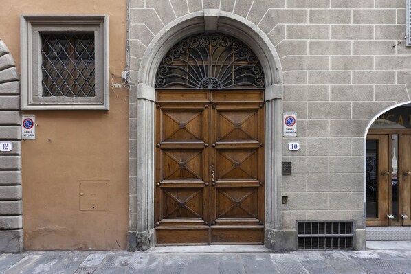 Puerta doble de madera con un arco metálico decorativo encima, ubicada en un edificio de piedra con letreros numerados que indican su ubicación.