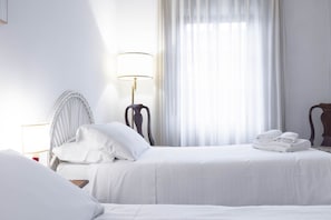 Habitación de hotel con dos camas, una mesita de noche con lámpara y una ventana con cortinas traslúcidas.