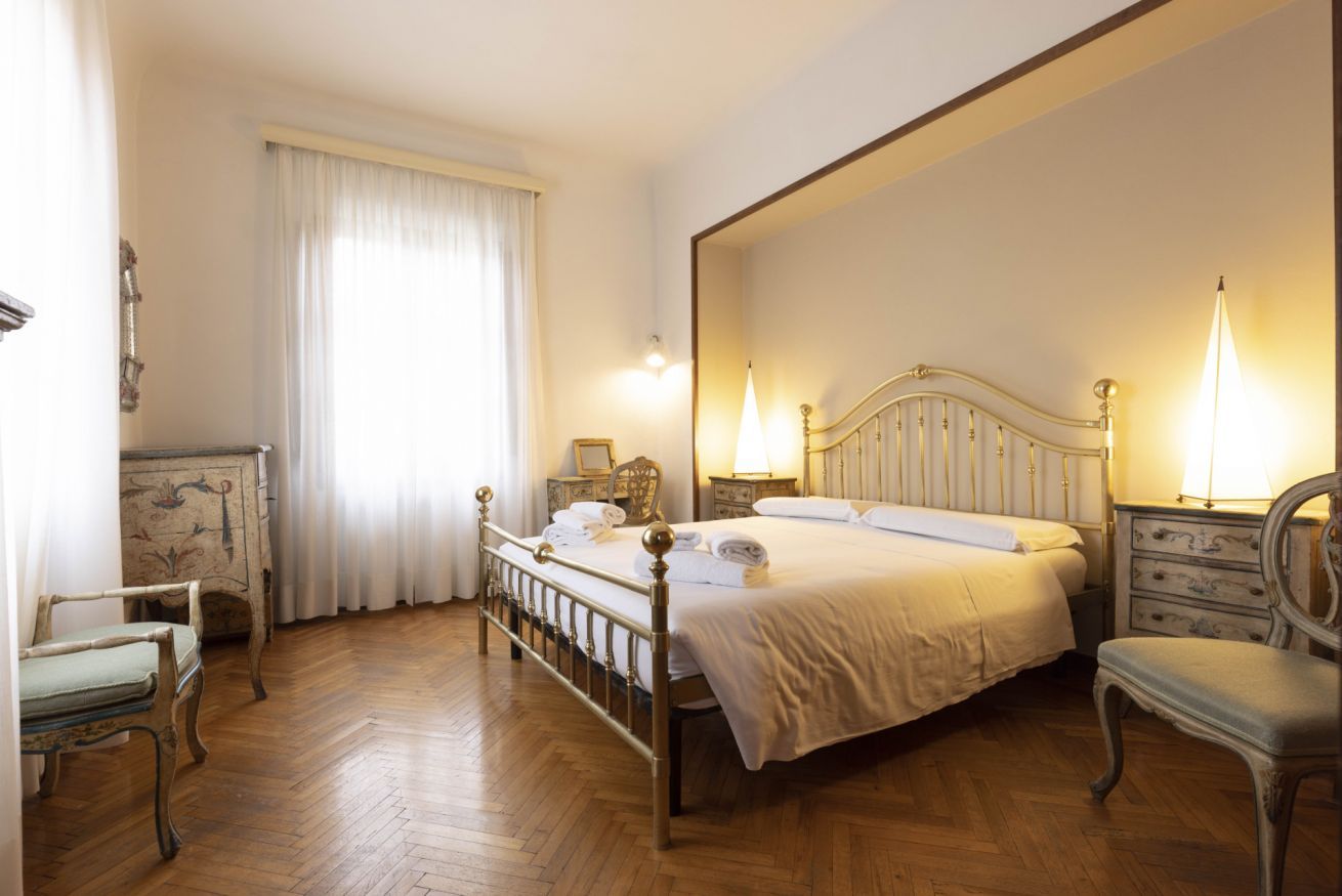 A bedroom with a wooden floor, a bed with white linens, two bedside tables with lamps, and a chair.