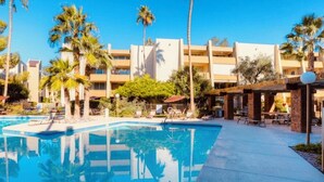 Um hotel com piscina, palmeiras e um pátio com espreguiçadeiras.