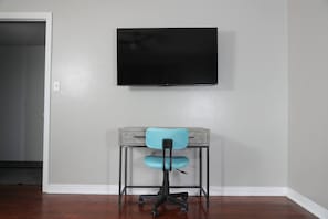 A room with a flat-screen TV mounted on the wall, a desk with a chair, and a wooden floor.