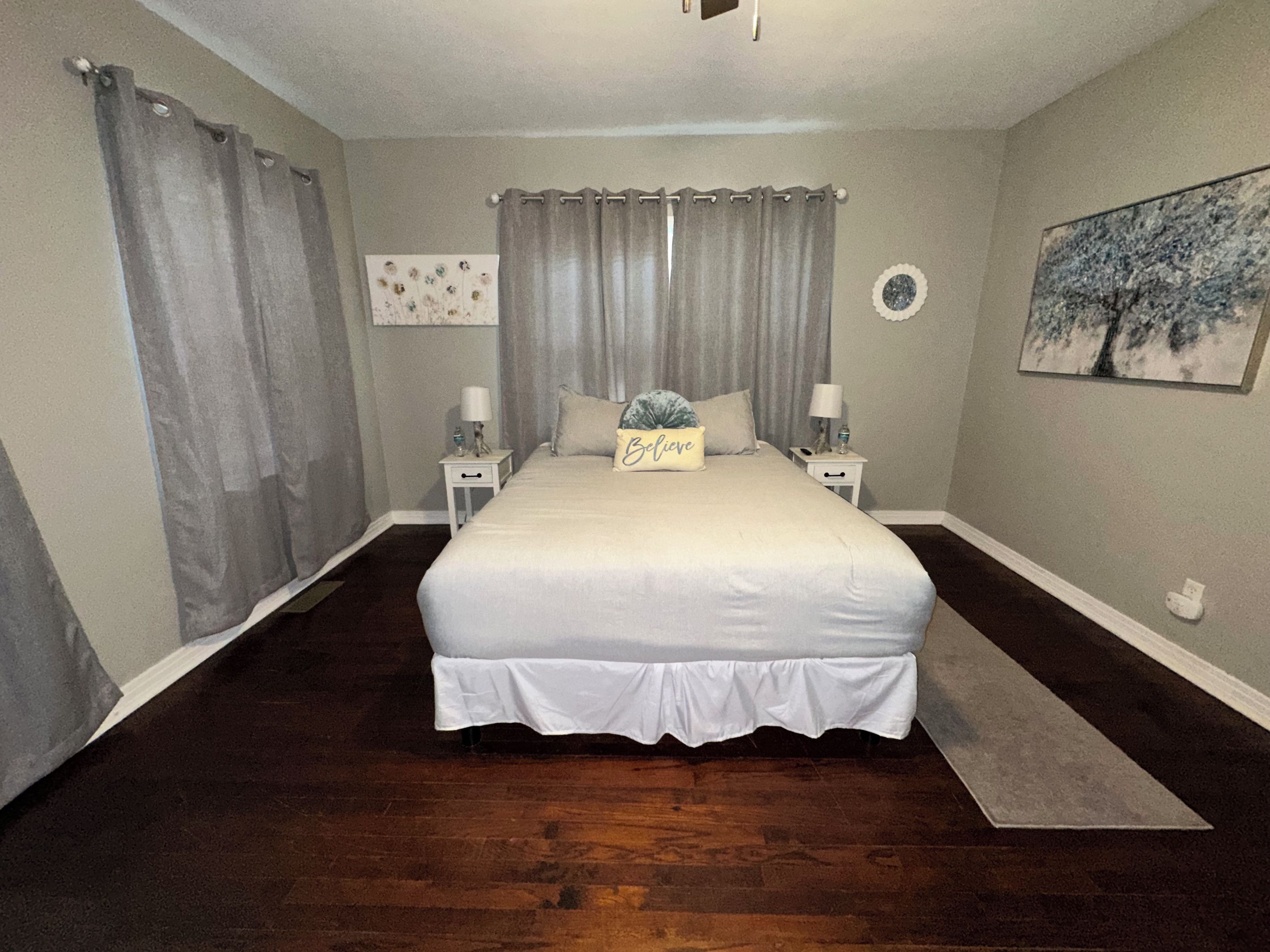 A bedroom with a large bed, two bedside tables with lamps, a nightstand, a wall clock, and a painting.