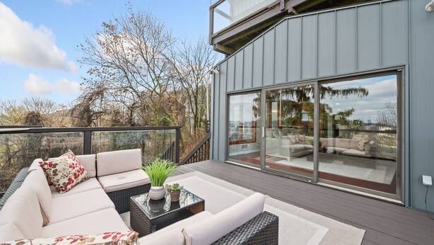 A modern outdoor seating area with a sofa and a table, a glass door leading to a balcony, and a view of trees and a building in the background. - Beautiful Hillside Home with Incredible City Views!! Great Location and Privacy! (Pittsburgh)
