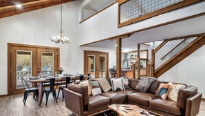 A spacious living room with a brown leather sectional sofa, a dining table with chairs, a chandelier, and a staircase leading to an upper level.