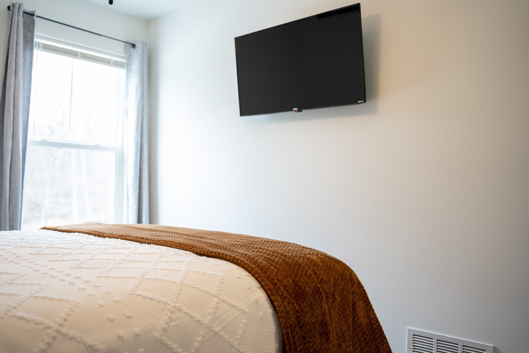 Ein Schlafzimmer mit einem Bett, einem Fenster mit Vorhängen, einem an der Wand montierten Fernseher und einem weißen Lüftungsgitter.
