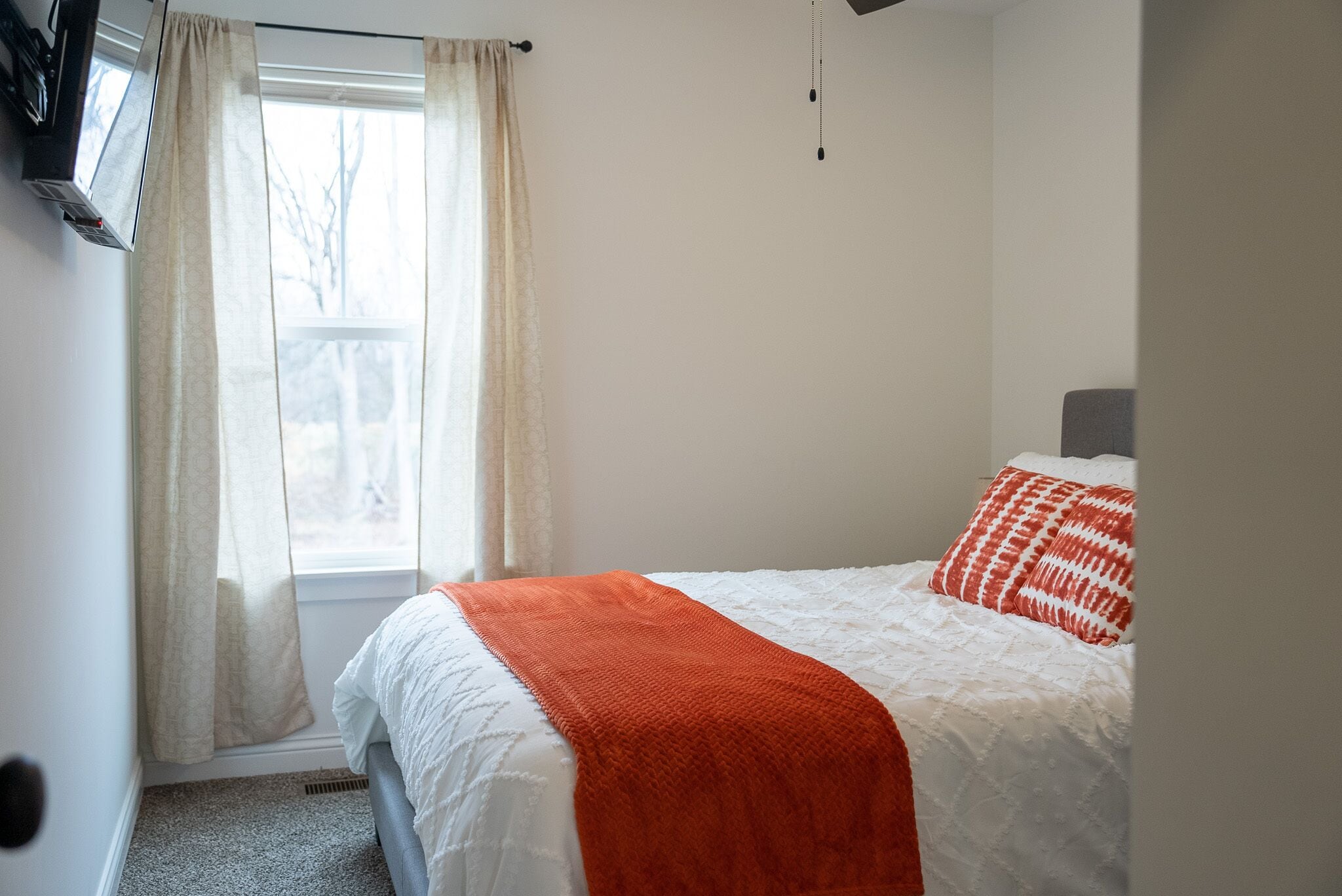 Un dormitorio con cama, ropa de cama blanca, una manta roja y una ventana con cortinas.