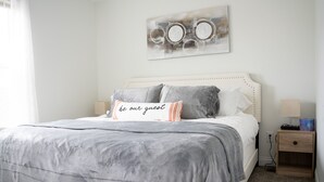 Una cama bien hecha con una paleta de colores gris y blanco, una mesita de noche con lámpara, y una obra de arte colgada en la pared sobre la cama.