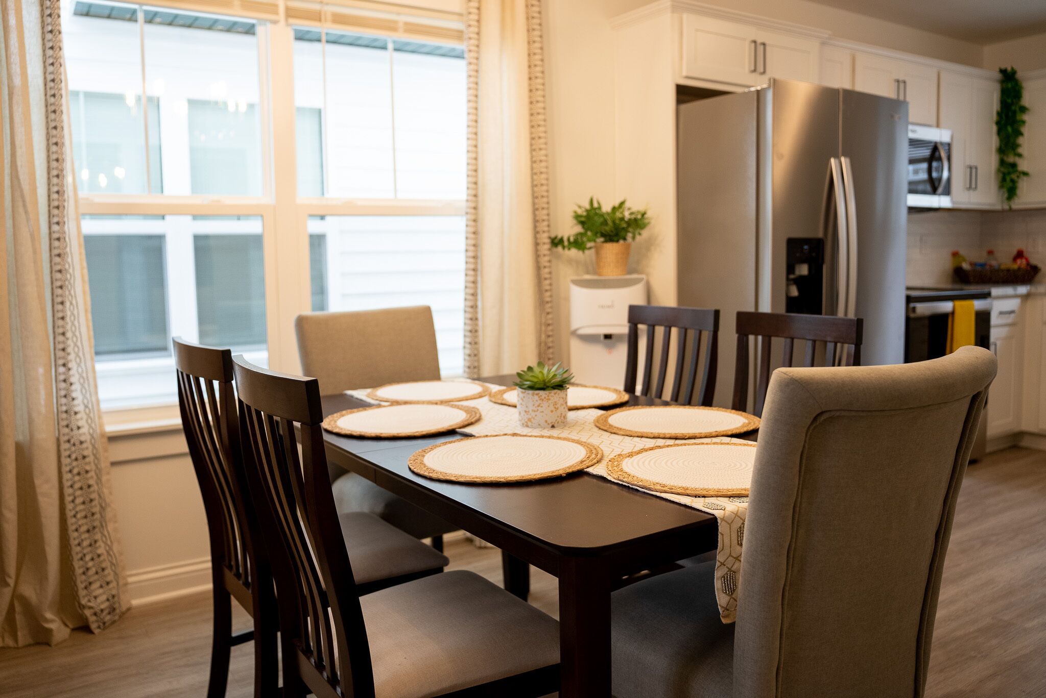 A dining table with six chairs, a central plant, and a kitchen area with appliances in the background.