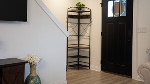 Rincón con una puerta negra, pared blanca, suelo de madera, estante metálico negro con una planta y un jarrón con flores blancas.