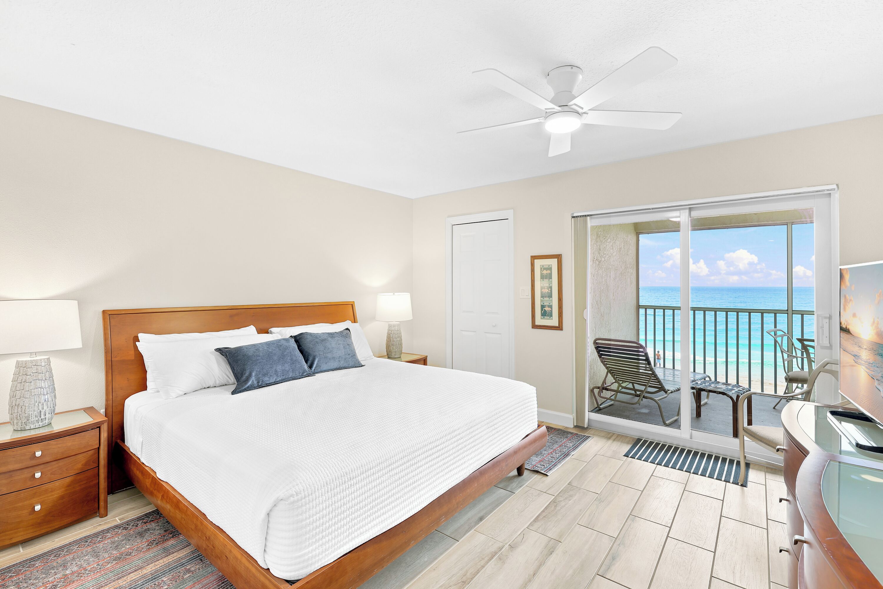 A hotel room with a bed, bedside lamps, a ceiling fan, a door, a framed picture, and a balcony with a view of the ocean.